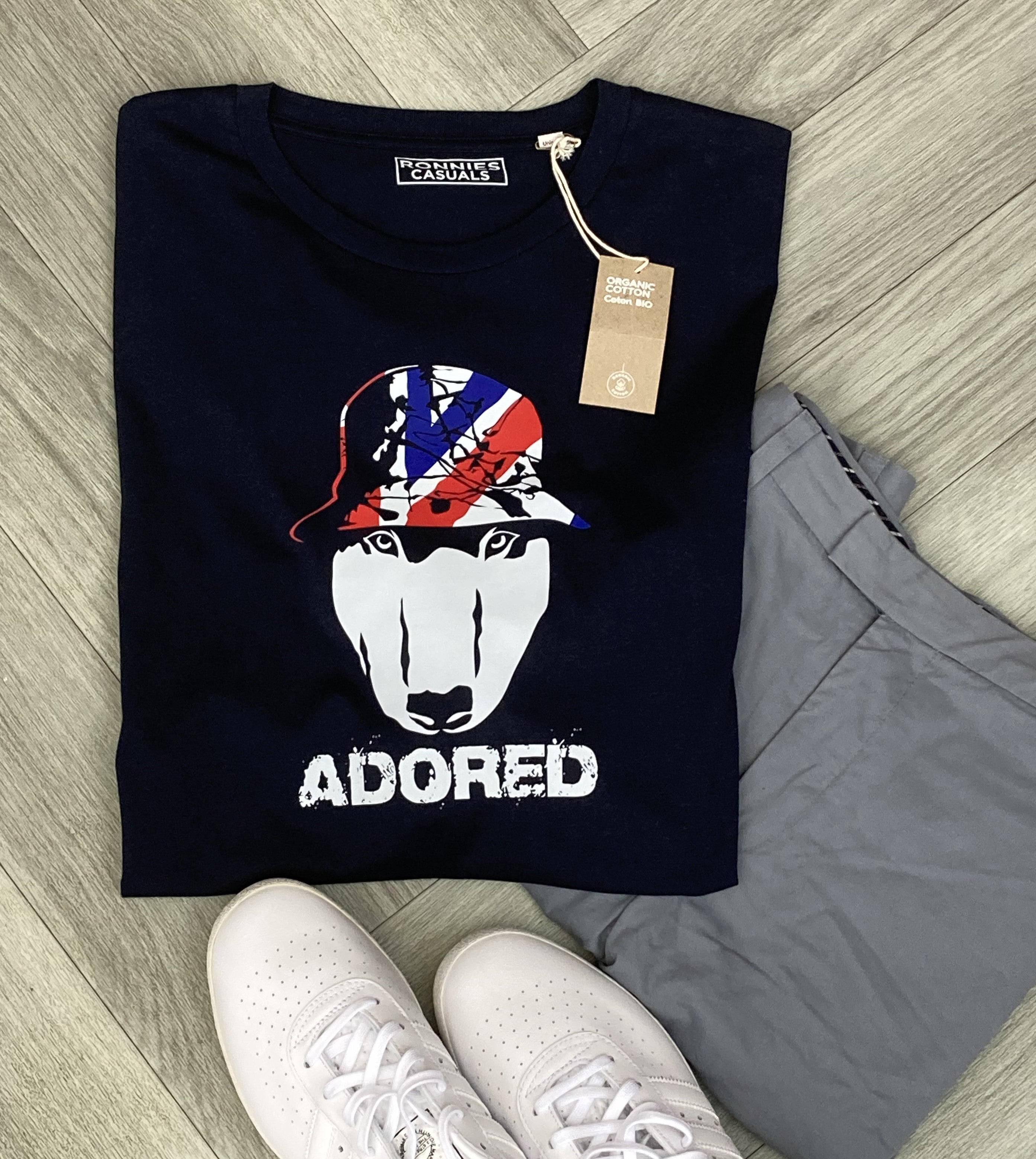 Black T-shirt printed with an English Bull Terrier Dog face wearing a bucket hat in red and blue and the word Adored underneath. Grey trousers and trainers laying on a grey floor.