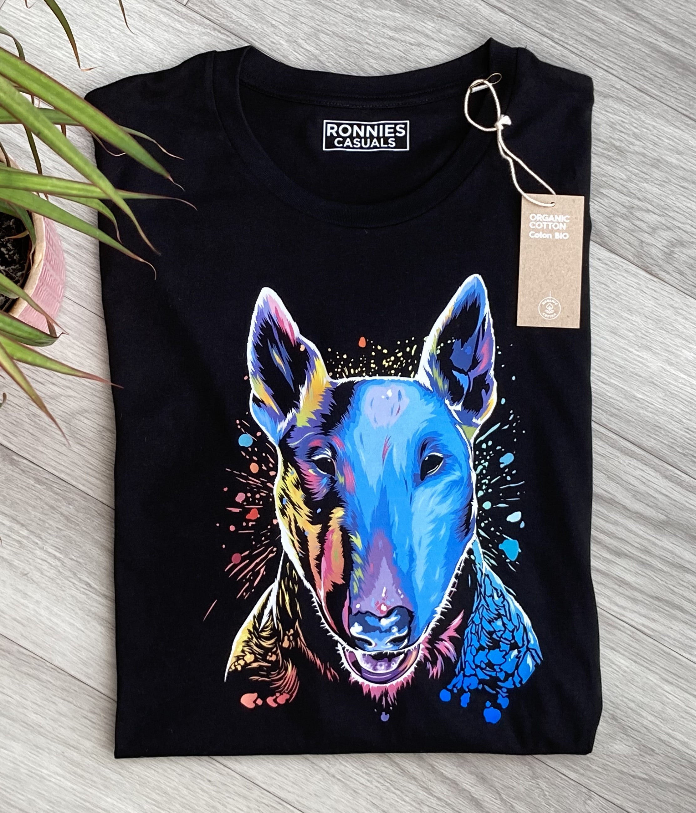 Black T-shirt printed with an English Bull Terrier Dog face in bright colours and paint splashes showing a clothes label, grey floor and plant
