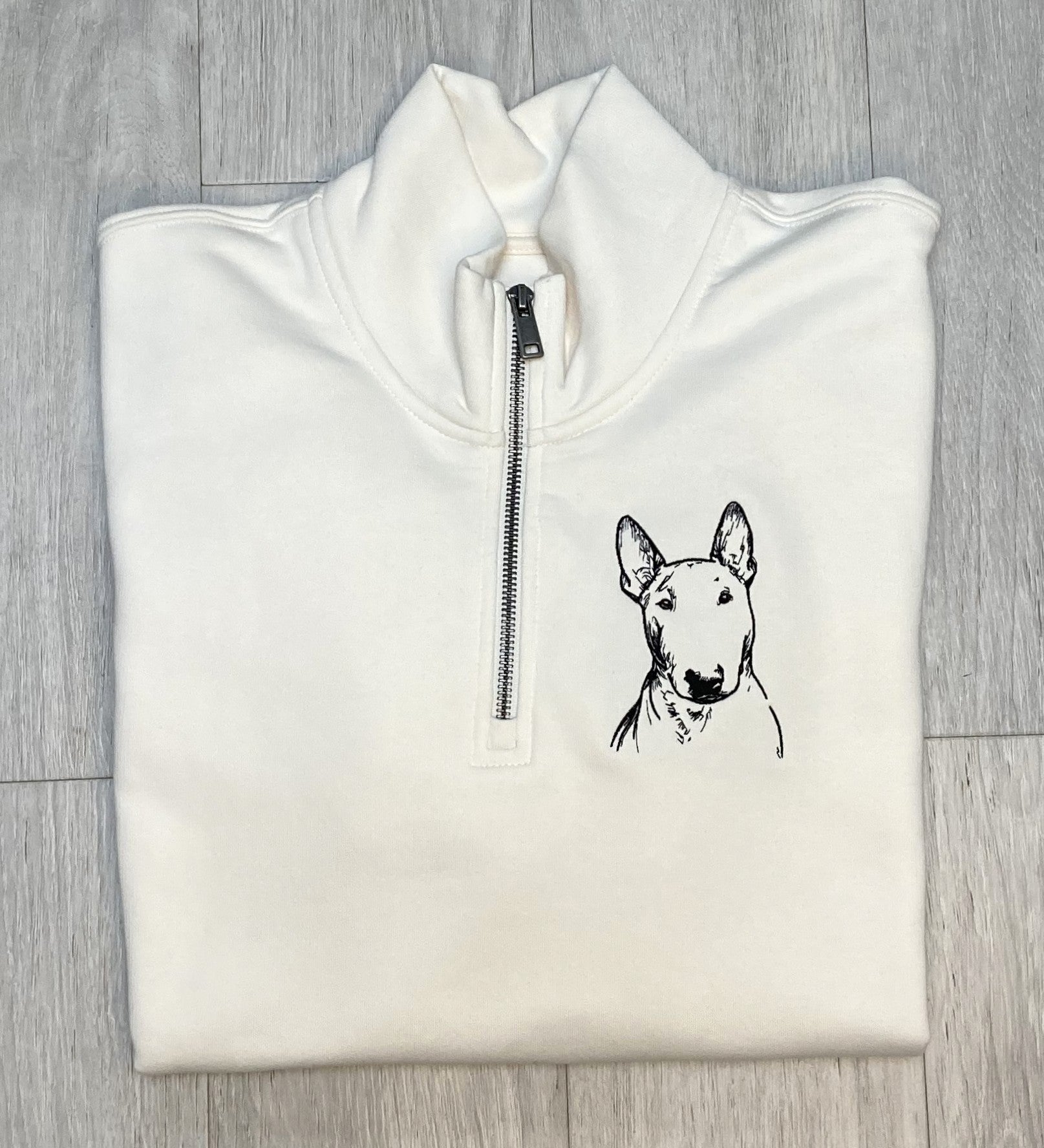 Ivory 1/4 zip sweatshirt
 with a bull terrier embroidered on the left breast laying on a wooden floor
