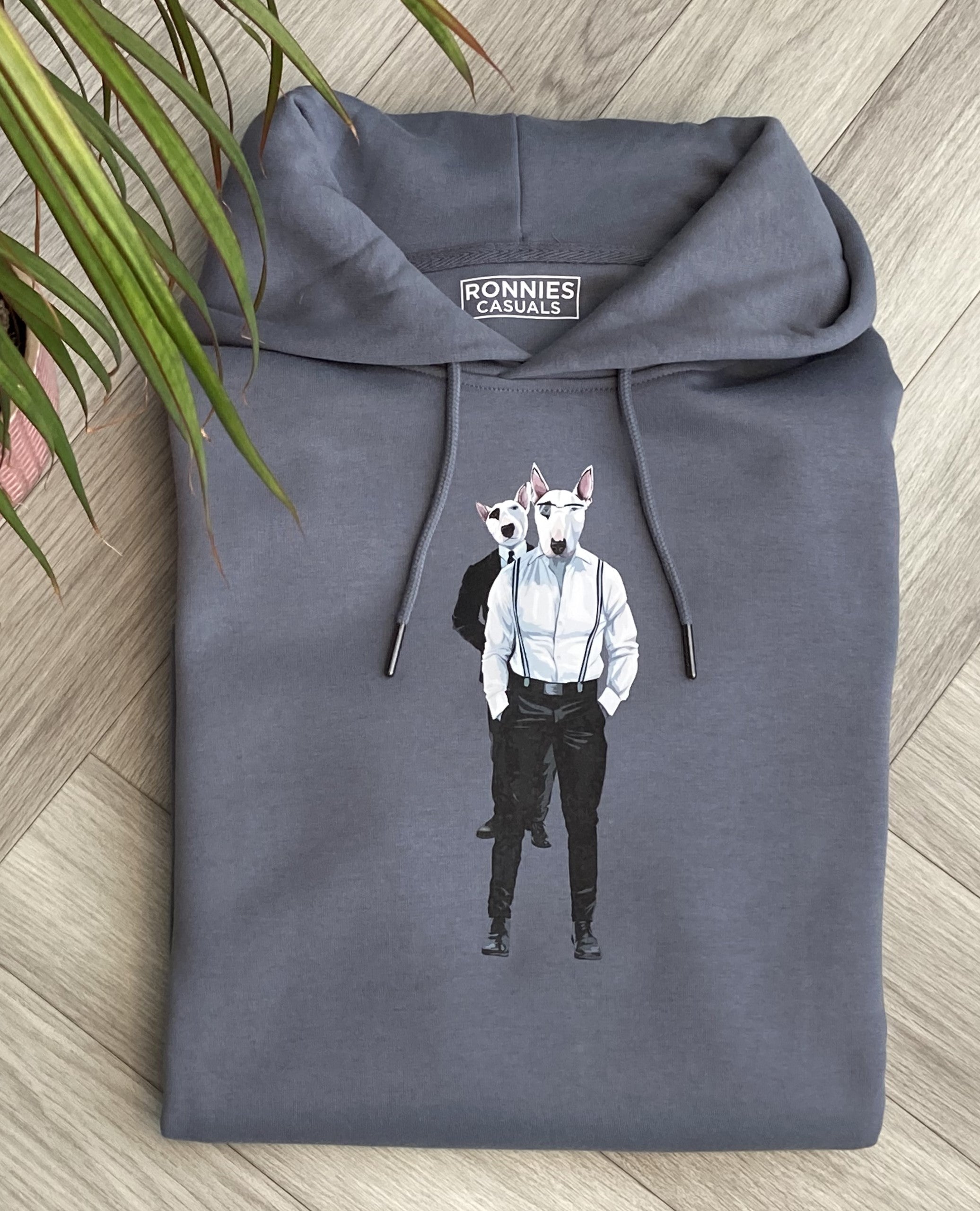 Mineral Grey Hoodie printed with English Bull terriers wearing suits and braces. Laying on a floor with plant leaves partially covering up one side.