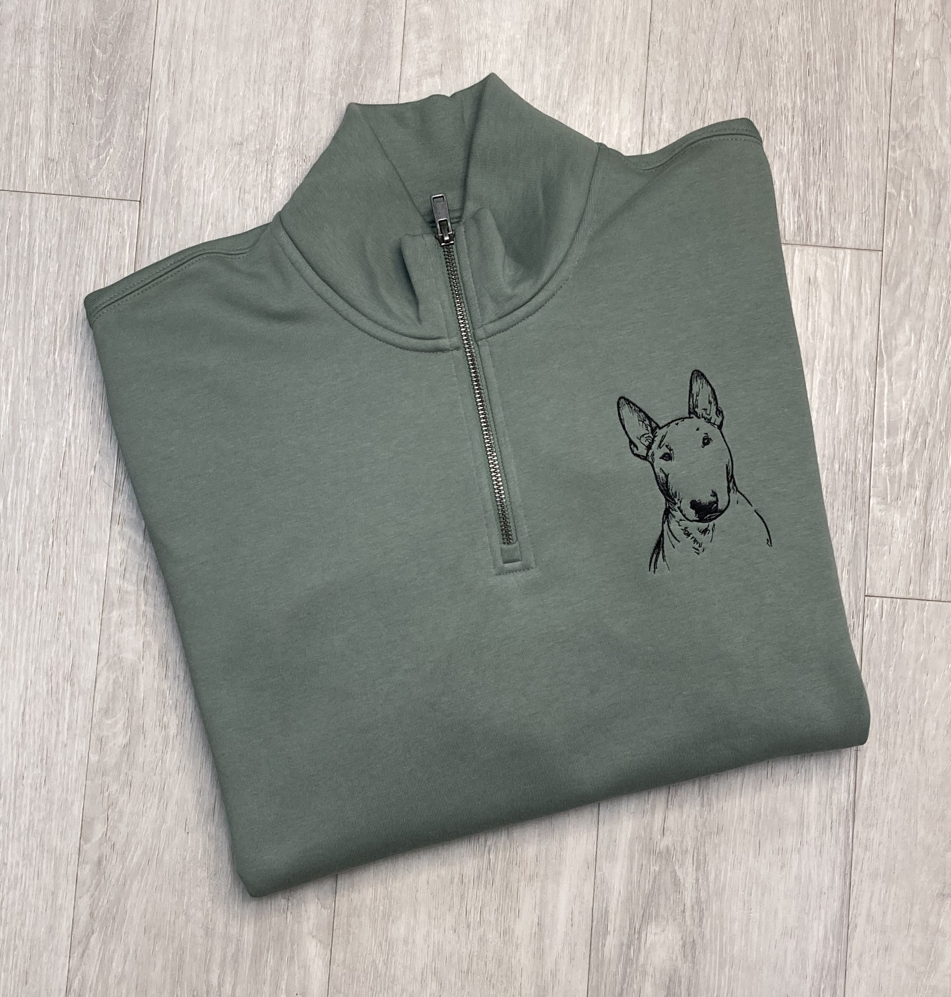 Folded green 1/4 zip neck sweatshirt with a english bull terrier embroidered on the left breast kaying on a wooden floor